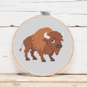 May include: A cross-stitch embroidery of a bison in shades of brown, tan, and white, set within a wooden embroidery hoop. The bison is depicted in a simple, geometric style, with a detailed outline. The background is white.