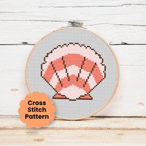 Puede incluir: Un bastidor de bordado circular de madera que contiene un patrón de punto de cruz de una concha marina. La concha está representada en tonos coral y rosa claro con contornos marrón oscuro sobre un fondo de tela gris claro. Una etiqueta color melocotón con bordes festoneados dice Cross Stitch Pattern.