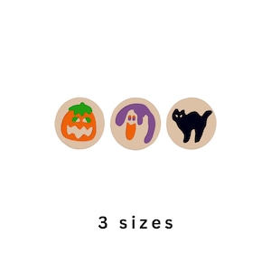 May include: Three circular beige buttons featuring Halloween-themed designs: an orange jack-o'-lantern with a green stem, a purple and orange ghost, and a black cat silhouette. The text 3 sizes is printed in black below the buttons.