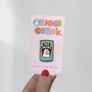 May include: A metal enamel needle minder pin shaped like a blue food can with a black label featuring the text TOE BEANS and a white cat paw print. The pin is attached to a light pink backing card with the text STITCH CHICK and NEEDLE MINDER.