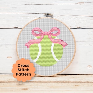 May include: A circular wooden embroidery hoop containing a cross stitch pattern of a lime green tennis ball with white curved lines, topped with a pink bow. An orange scalloped label in the bottom left corner displays the text Cross Stitch Pattern.