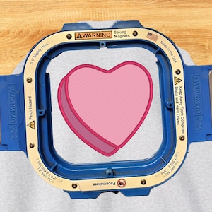 以下が含まれることがあります： 14cmの青とベージュのMighty Hoop刺繍枠。ライトグレーの生地にピンクのハート模様が施されている。枠には警告ラベル、Strong Magnets、Made in the USAの文字、特許番号が印刷されている。