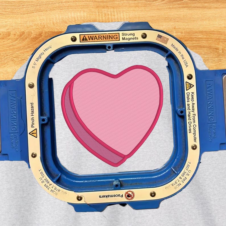 以下が含まれることがあります： 14cmの青とベージュのMighty Hoop刺繍枠。ライトグレーの生地にピンクのハート模様が施されている。枠には警告ラベル、Strong Magnets、Made in the USAの文字、特許番号が印刷されている。