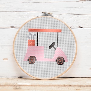 Könnte beinhalten: Ein Kreuzstich-Stickrahmen mit einem rosa Golfwagen-Design. Der Golfwagen hat schwarze Räder und ein Lenkrad, mit einem korallenfarbenen Dach und Akzenten. Der Hintergrund ist weiß und der Rahmen ist aus Holz.