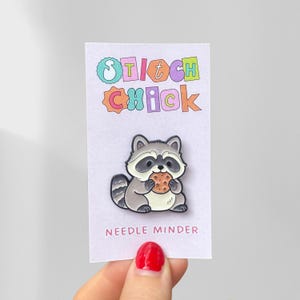 May include: A metal enamel pin featuring a grey and white raccoon holding a brown chocolate chip cookie. The pin is attached to a white backing card with the text Stitch Chick at the top and the words NEEDLE MINDER printed at the bottom.