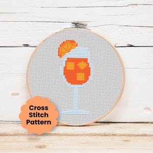 Könnte beinhalten: Ein Kreuzstichmuster in einem hölzernen Stickrahmen, das ein Cocktailglas mit einer orangefarbenen Flüssigkeit, Eiswürfeln und einer Orangenscheibe als Garnitur zeigt. Ein gewelltes orangefarbenes Etikett unten links zeigt den Text Cross Stitch Pattern.