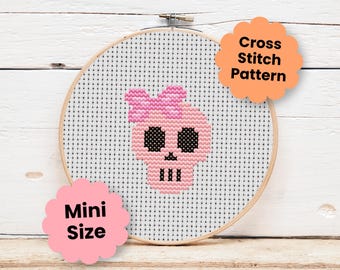 May include: A round wooden embroidery hoop containing white Aida fabric with a cross stitch design of a light pink skull wearing a pink bow. The skull features black eyes and a black nose and mouth. Text on the image reads Cross Stitch Pattern and Mini Size.