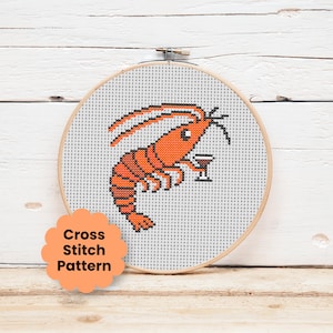 May include: A cross stitch pattern in a circular wooden embroidery hoop featuring an orange shrimp holding a small cocktail glass. The design is stitched on white Aida fabric with a scalloped peach-colored label reading Cross Stitch Pattern.