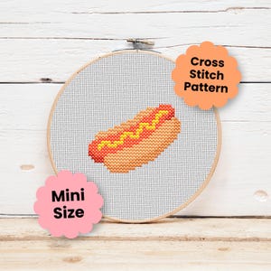 May include: A round wooden embroidery hoop containing white Aida fabric with a cross stitch pattern of a hot dog with a yellow mustard line. An orange scalloped label reads Cross Stitch Pattern and a pink scalloped label reads Mini Size.