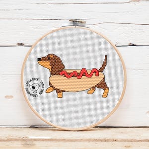 Op de afbeelding: Een kruissteek borduurring met een teckel in een hotdogbroodje. De hond is bruin en bruin, met een rode hotdog. De tekst "Stitch Chick" staat aan de linkerkant van de ring.
