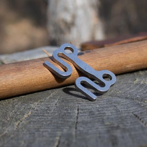 Może przedstawiać: Para małych, srebrnych metalowych haczyków o unikalnym, zakrzywionym kształcie. Haczyki umieszczone są na drewnianym kołku, spoczywającym na zwietrzałej, szarej drewnianej powierzchni. Haczyki mają pętlę na jednym końcu i zakrzywiony kształt haka.