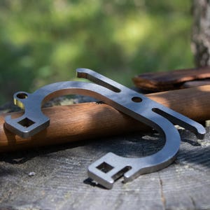 Pode incluir: Uma ferramenta multiuso prateada com um design curvo, apresentando uma abertura quadrada e uma extremidade ranhurada. Descansa sobre uma superfície de madeira, sugerindo uso ao ar livre. O design da ferramenta implica que é para várias tarefas.