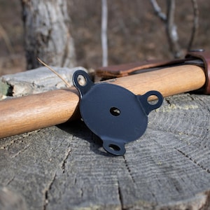 Puede incluir: Un protector de cabeza de hacha de metal negro con tres puntos de fijación circulares. El protector descansa sobre un mango de hacha de madera y un tocón de árbol desgastado. El mango del hacha es de madera clara.