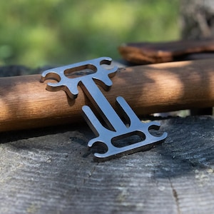 Könnte beinhalten: Ein silberfarbenes Multitool liegt auf einem Holzdübel, vor einem verschwommenen natürlichen Hintergrund. Das Werkzeug hat ein einzigartiges, offenes Design mit mehreren ausgeschnittenen Formen und einem Mittelbalken. Der Dübel ist etwa 30 cm lang.