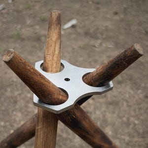 Può includere: Una piastra di collegamento per treppiede in metallo color argento con tre aperture circolari, che sostiene tre bastoni di legno cilindrici in una configurazione divaricata. La piastra metallica presenta un piccolo foro centrale e una finitura opaca.