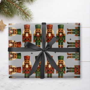 May include: Gray wrapping paper featuring a repeating pattern of toy nutcrackers in red and green uniforms. The wrapped gift is tied with a black ribbon. A roll of matching paper is visible to the right. The scene is decorated with pine branches, pine cones, and star-shaped ornaments.