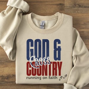 Puede incluir: Una sudadera beige con las palabras "GOD & CROSS COUNTRY" en letras de bloque azules y rojas. La palabra "Cross" está en cursiva. La frase "running on faith" está debajo, con dos pequeñas huellas.