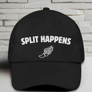 May include: Black baseball cap featuring the white text "SPLIT HAPPENS" and a white winged foot graphic. The cap has a curved peak and a button on top. The hat is made of a soft material.