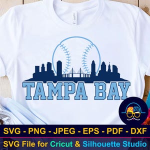 Può includere: T-shirt bianca con un design di baseball blu e la scritta "TAMPA BAY". Il design include una palla da baseball, uno skyline della città e la scritta "TAMPA BAY" in grassetto. La maglietta è un file SVG per Cricut & Silhouette Studio.