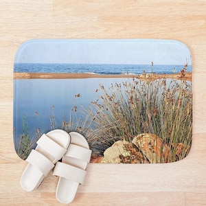Könnte beinhalten: Eine Badematte mit einem Foto einer Strandszene. Das Foto zeigt einen Sandstrand mit blauem Wasser und eine grasbewachsene Fläche mit Steinen. Das Foto ist aus einer niedrigen Perspektive aufgenommen und zeigt auf den Strand.