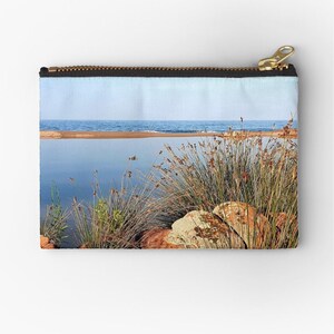 Könnte beinhalten: Rechteckige Reißverschlusstasche mit Landschaftsmotiv. Das Design zeigt ein ruhiges Gewässer, einen Sandstrand und einen klaren blauen Himmel. Hohes Gras und Felsen befinden sich im Vordergrund, mit einem goldenen Reißverschluss.