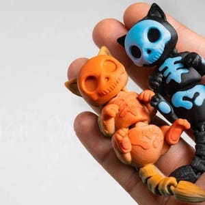May include: Two articulated cat figurines. One is orange with a skull face, the other is black with a blue skull face and blue skeleton markings. The figurines are held in a hand.