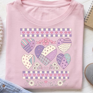 May include: A light pink t-shirt featuring a patchwork heart design. The design includes six hearts in various patterns and colors, with floral and ribbon accents. The top and bottom of the design have a border of hearts and checkered patterns.