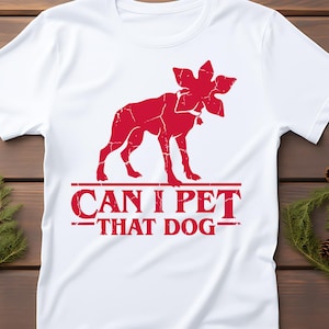 May include: White t-shirt featuring a red graphic of a dog-like creature with a flower-like head. The text "CAN I PET THAT DOG" is printed in red below the graphic. The shirt is made of a soft material.