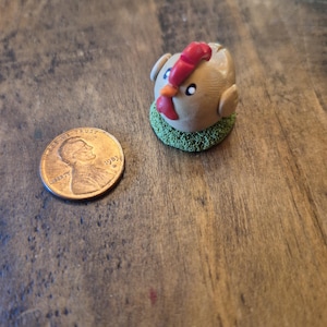 May include: A small ceramic figurine of a brown chicken with a red comb and a green base. The chicken is sitting on a green base that looks like grass.