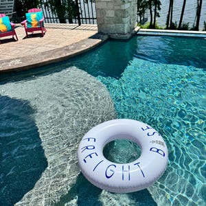 Könnte beinhalten: Ein weißer aufblasbarer Schwimmring mit dem blauen Schriftzug "FREIGHT BASE" auf der Oberseite, der im klaren blauen Wasser eines Swimmingpools treibt. Der runde Ring zeigt ein kleines grafisches Leuchtturm-Symbol über dem Text.