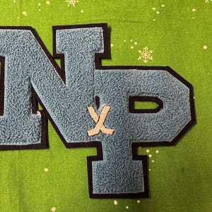 May include: A light blue felt letter patch with the letters "NP" in a varsity style, outlined in black. A small white hockey stick design is centered between the letters. The patch is on a green fabric background with white snowflake accents.