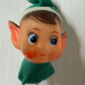 May include: A vintage elf head on a stick, featuring a smiling face with blue eyes, rosy cheeks, and brown hair. The elf wears a green felt hat with a white brim and a small gold bell. The elf has large, pointed ears and a green felt collar.