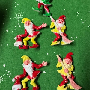 May include: A collection of colorful Christmas elf figurines. The elves are depicted in various poses, wearing red, yellow, and green outfits with white beards and hats. Some hold gifts or brooms. The background is a green fabric with white snowflake patterns.