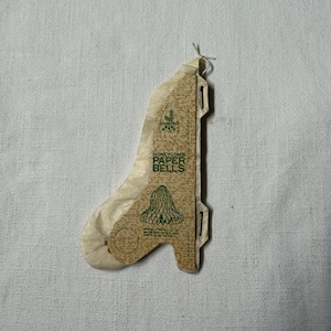 Puede incluir: Una campana de papel de nido de abeja vintage, en beige y verde. El embalaje dice "HONEYCOMB PAPER BELLS" y muestra un gráfico de campana. La campana está plegada, lista para montar.