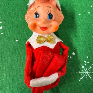 Peut inclure: Figurine de lutin de Noël vintage avec un visage souriant, des yeux bleus et un nœud papillon doré. Le lutin porte une tenue rouge avec des bordures blanches et un chapeau blanc. Le fond est un tissu vert avec des motifs de flocons de neige blancs.