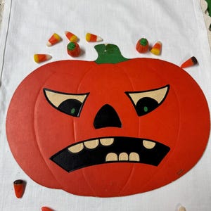 May include: A vintage orange paper pumpkin decoration with a black mouth and eyes, and a green stem. Candy corn and small pumpkin candies are scattered around the pumpkin. A festive Halloween decoration.