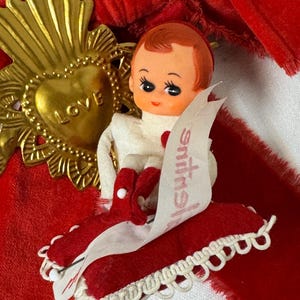 Puede incluir: Adorno vintage del Día de San Valentín con una muñeca de pelo rojo, vestida de blanco y con zapatos rojos. La muñeca está sentada sobre un cojín de terciopelo rojo con un ribete de encaje blanco. También se ve un corazón dorado con la palabra "LOVE" y una cinta de terciopelo rojo.