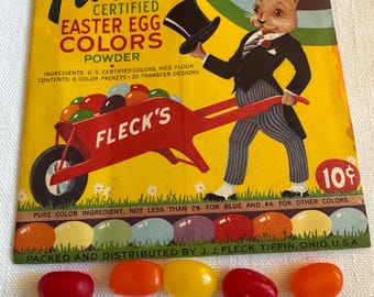 May include: A vintage Fleck's Easter Egg Colors powder box featuring a rabbit in a tuxedo and a wheelbarrow of eggs. Six jelly beans in red, orange, and yellow are in front of the box. The box is yellow.
