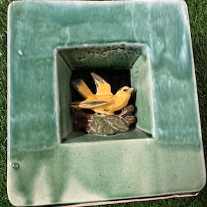 May include: A square, ceramic decorative piece with a teal green frame. Inside the frame is a recessed area featuring a yellow bird perched on a brown branch with green leaves. The bird has black accents. The piece is likely for home decor.