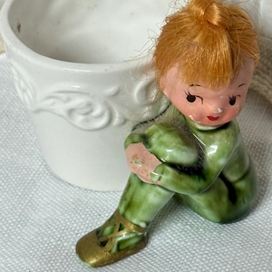 May include: A white ceramic planter with embossed floral details, accompanied by a seated figurine. The figurine has red hair, a green outfit, and gold shoes. The figurine is hugging its knees.