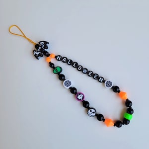 May include: A beaded Halloween-themed phone charm. The charm features black beads, orange geometric beads, and beads with Halloween-themed graphics. The word "HALLOWEEN" is spelled out in white letter beads. A black bat charm is at the top.