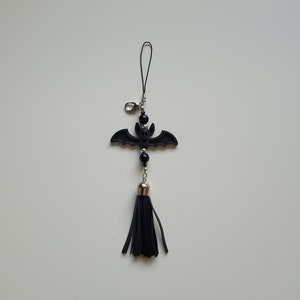 May include: Black bat charm with a black tassel hanging from a silver ring. The bat has a small face and is made of a textured material.