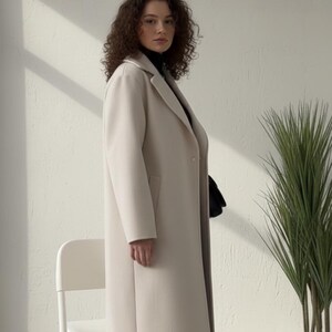 Può includere: Una donna indossa un lungo cappotto beige chiaro sopra un dolcevita nero e pantaloni neri. Il cappotto ha un design classico con chiusura a bottoni e tasche. Una sedia bianca e una pianta in vaso sono sullo sfondo.