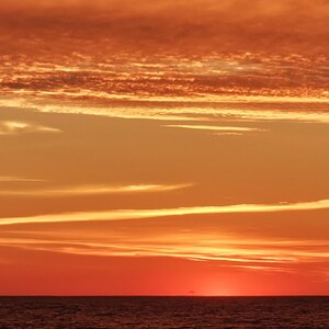 May include: A scenic view of a sunset over the ocean. The sky is filled with shades of orange, red, and yellow, with wispy clouds. The water is dark and calm, reflecting the colors of the sky. The overall scene is peaceful and serene.