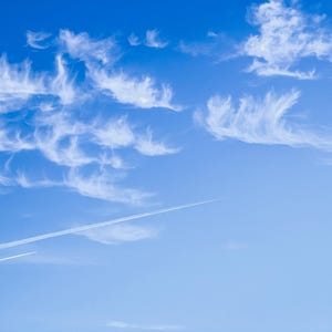May include: A bright blue sky filled with wispy white clouds. The clouds have a feathery appearance, with some appearing to be in the shape of brushstrokes. Two white contrails streak across the sky, adding a sense of movement.