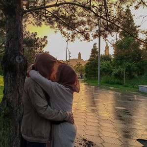 May include: A couple embraces under a tree in a park, with a building in the background. The woman is wearing a brown headscarf and the man is wearing a brown jacket.