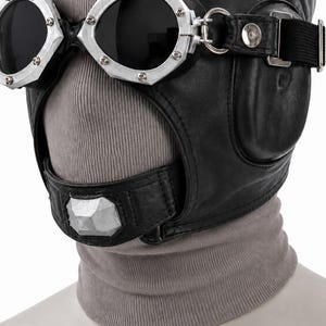 May include: A black leather aviator-style headgear featuring integrated dark-tinted goggles with silver-toned metal frames and rivets. The headgear includes a chin strap with a central silver-toned geometric accent and is worn over a grey ribbed fabric.