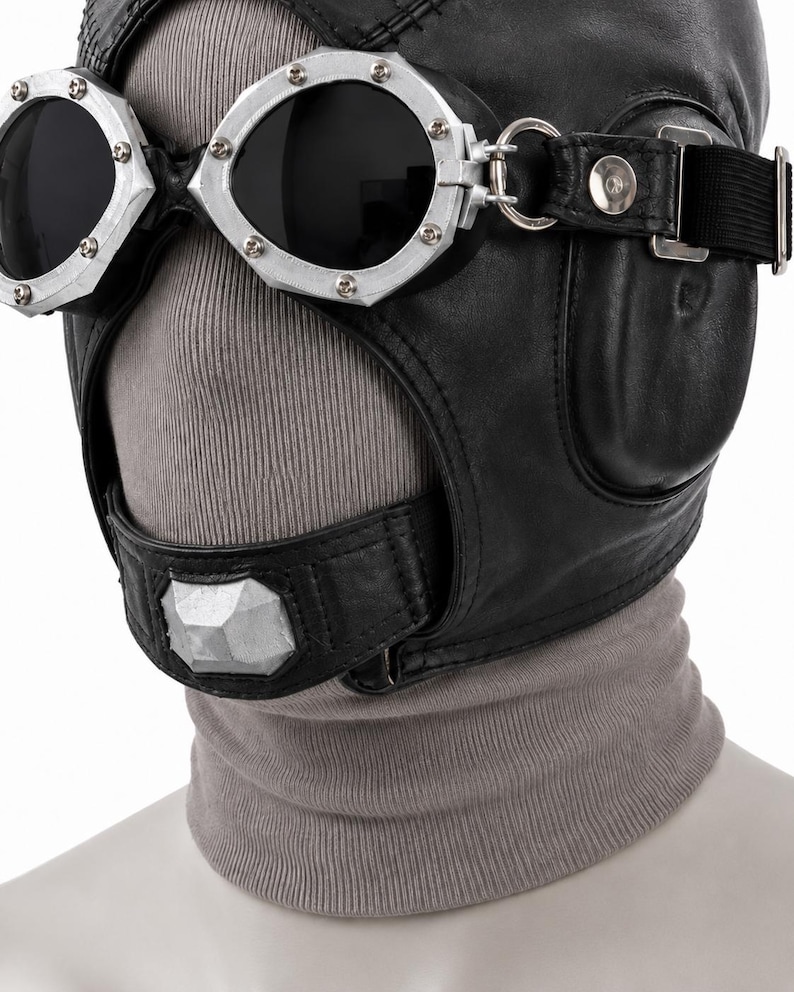 May include: A black leather aviator-style headgear featuring integrated dark-tinted goggles with silver-toned metal frames and rivets. The headgear includes a chin strap with a central silver-toned geometric accent and is worn over a grey ribbed fabric.
