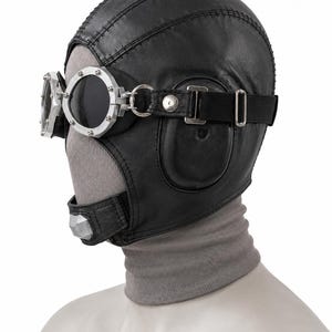 May include: A black leather aviator-style cap featuring stitched paneling and integrated ear flaps. The cap is paired with silver-toned metal goggles that have dark, circular lenses, secured by a black elastic strap and metal hardware.