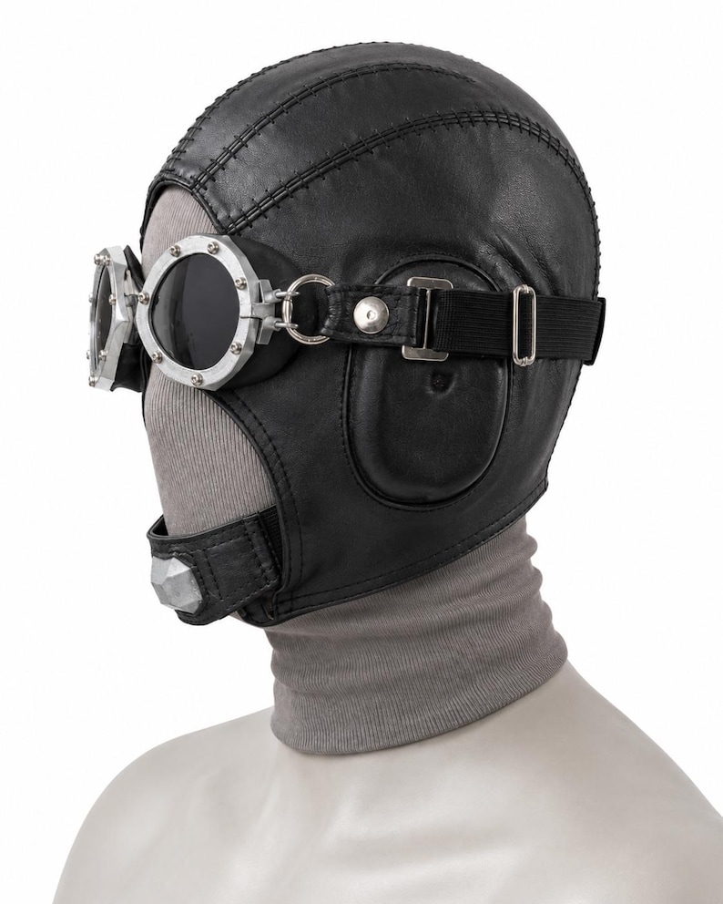 May include: A black leather aviator-style cap featuring stitched paneling and integrated ear flaps. The cap is paired with silver-toned metal goggles that have dark, circular lenses, secured by a black elastic strap and metal hardware.